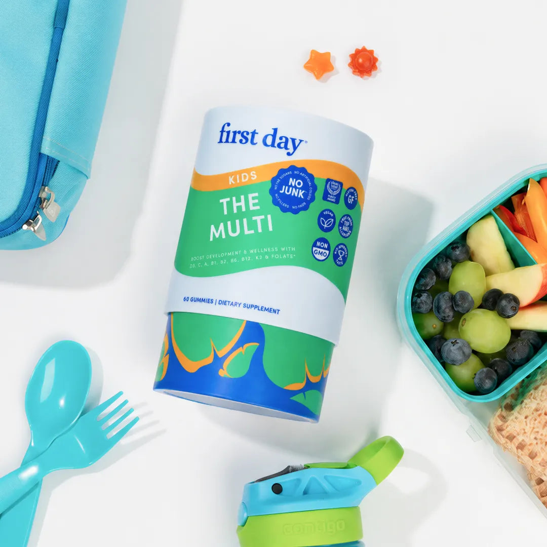 A First Day Kids 'The Multi' dietary supplement canister is displayed in a styled flat-lay on a white surface, surrounded by school and snack props including a blue lunch bag, utensils, and a fruit container.
