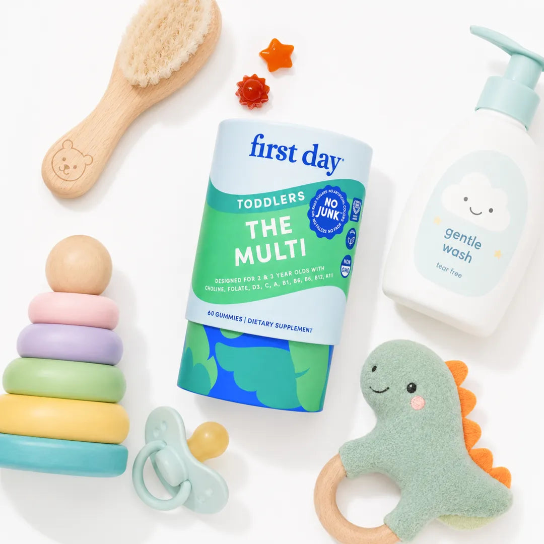 A styled flat-lay of baby and toddler products on a white background, featuring a container of First Day Toddlers 'The Multi' dietary supplement, a bottle of Gentle Wash soap, a wooden baby brush, a ring stack toy, a pacifier, and a dinosaur teether.
